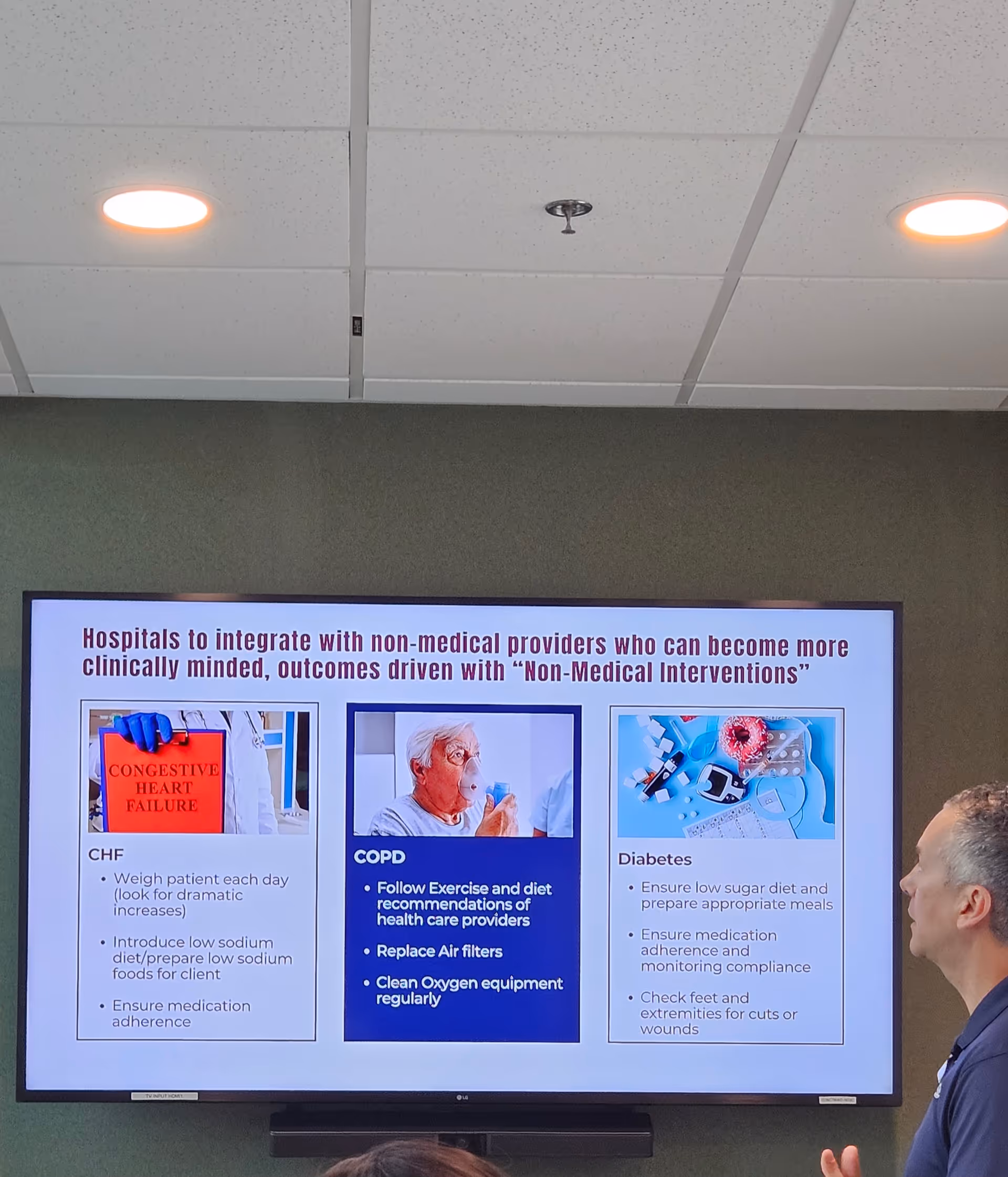 Indoor conference room with a large screen showing healthcare slides and a presenter.