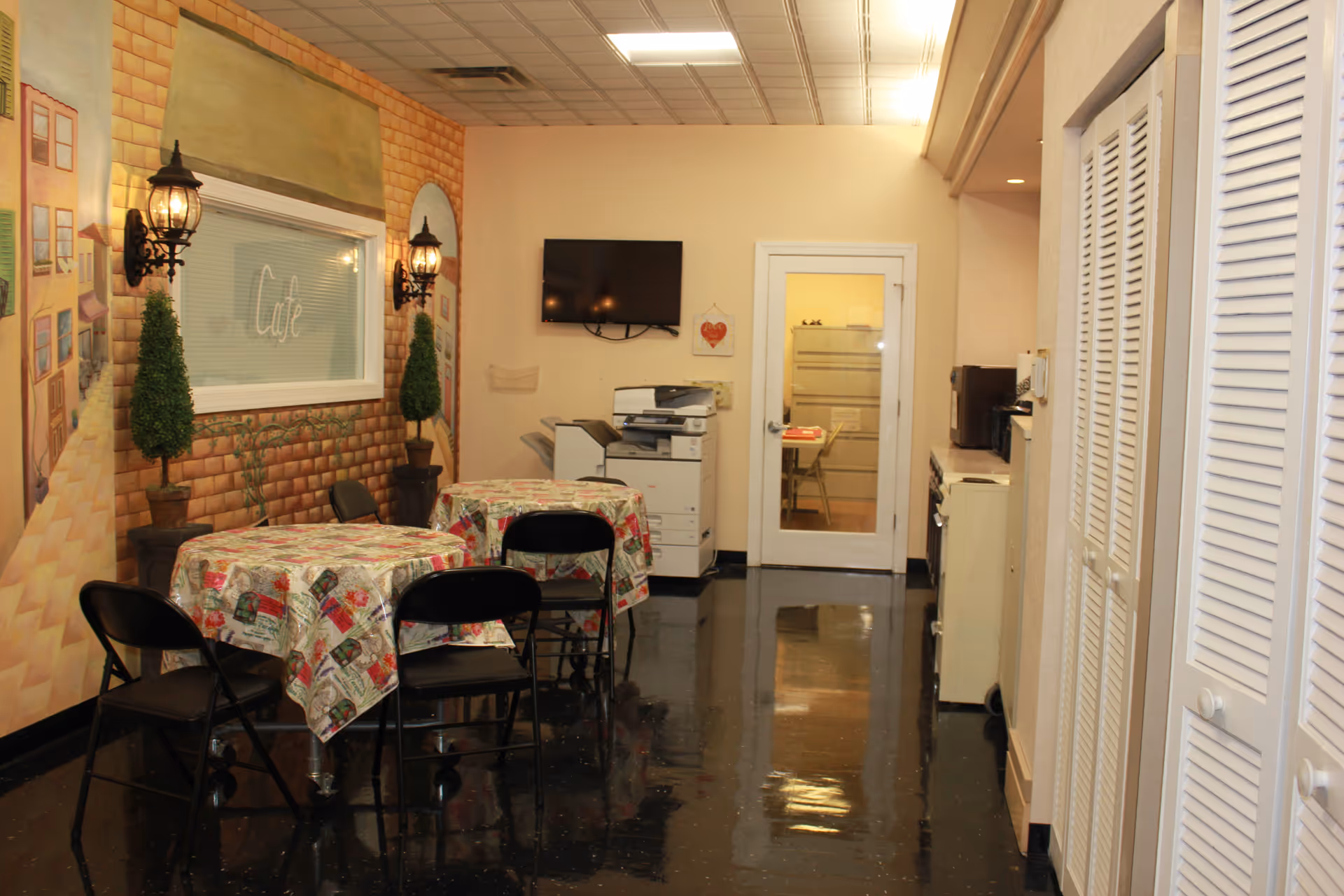 Indoor cafe style common area with round tables, tablecloths, folding chairs, and wall decor.