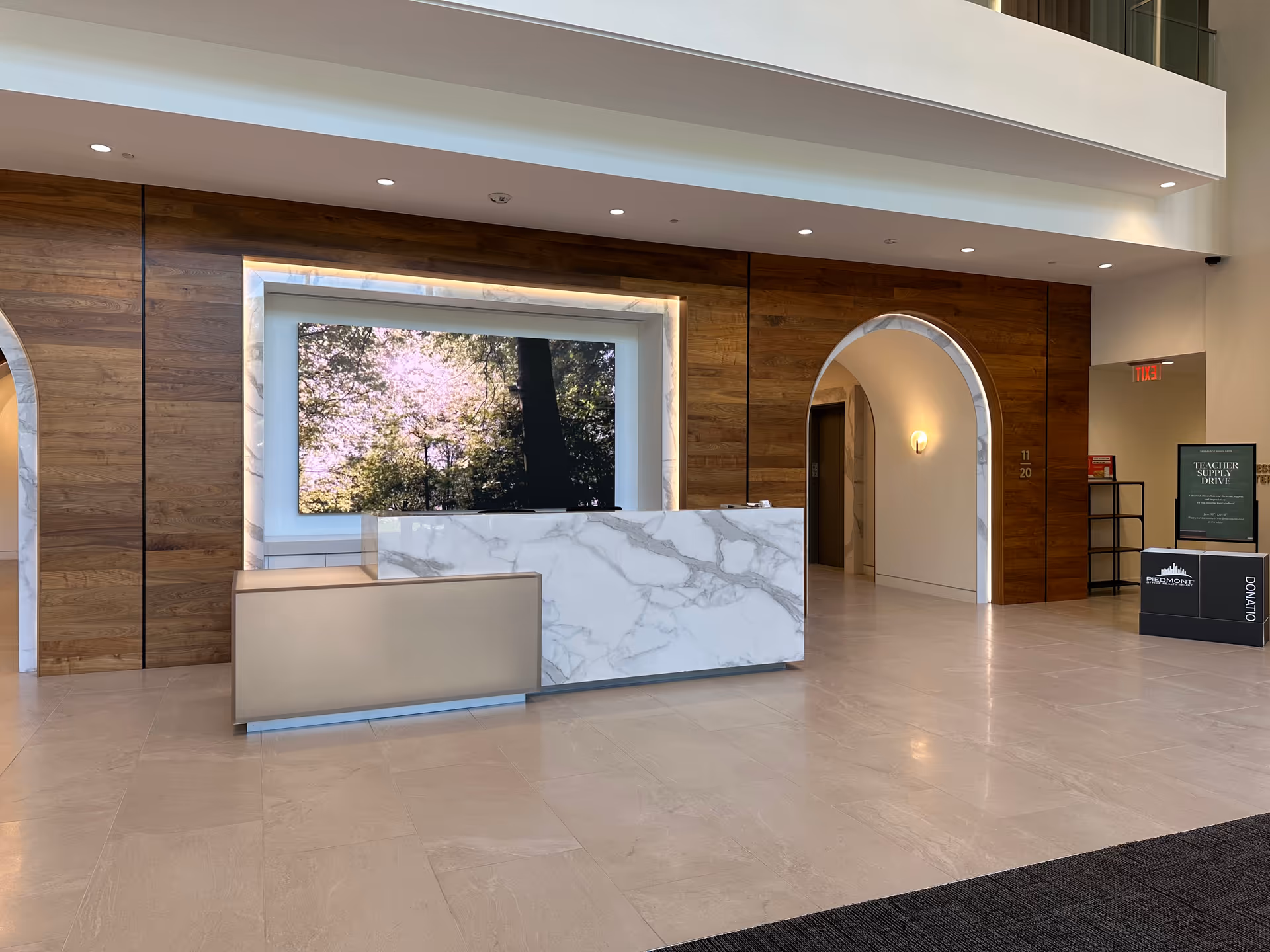 Modern lobby with marble reception desk, wood walls, arched doorway, and a nature scene on screen.