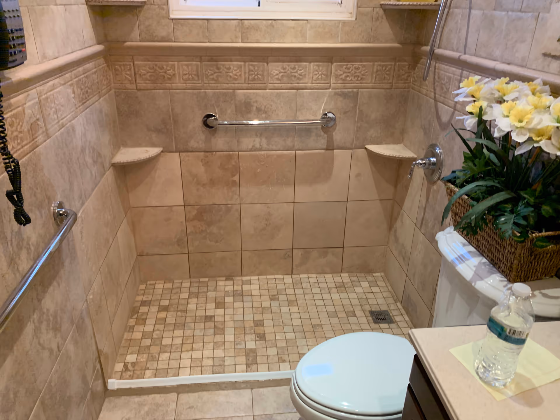 Accessible beige-tiled bathroom with grab bars, a walk-in shower, and a flower arrangement.