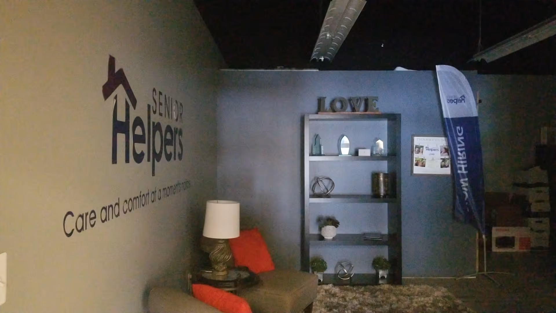 Interior lounge area featuring Senior Helpers logo wall, sofa, lamp, and decorative shelves.