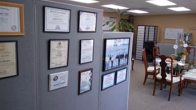 Partition wall displays framed certificates in a senior care facility; dining room chairs visible in background.