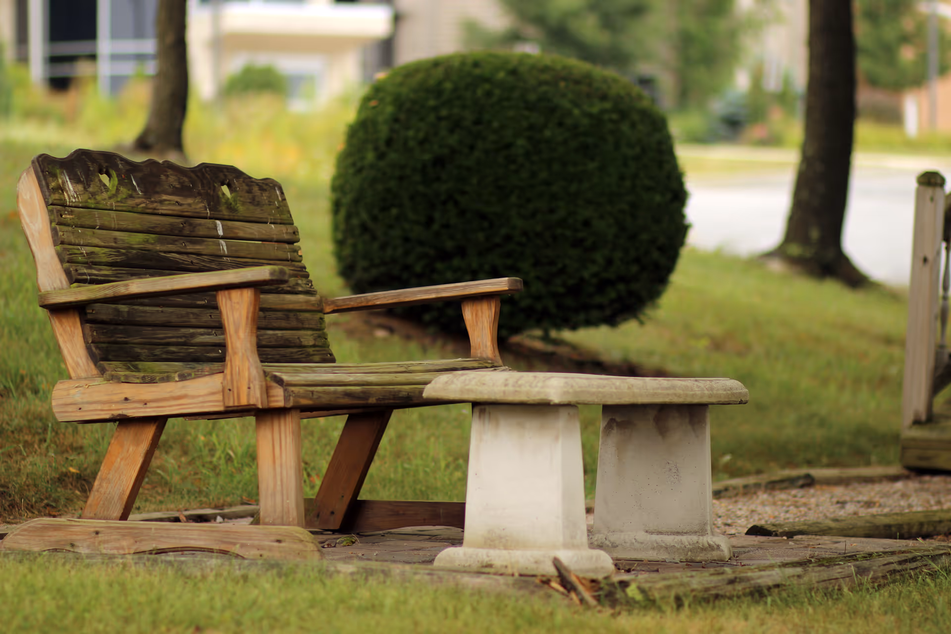 Weathered wooden bench and stone table on a grassy outdoor area with greenery.