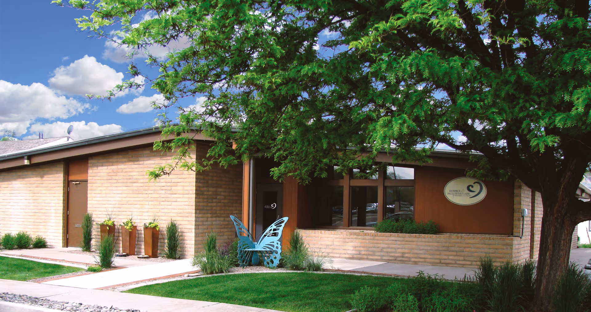 Brick exterior of HopeWest Administrative Offices with green lawn, tree, and butterfly sculpture.