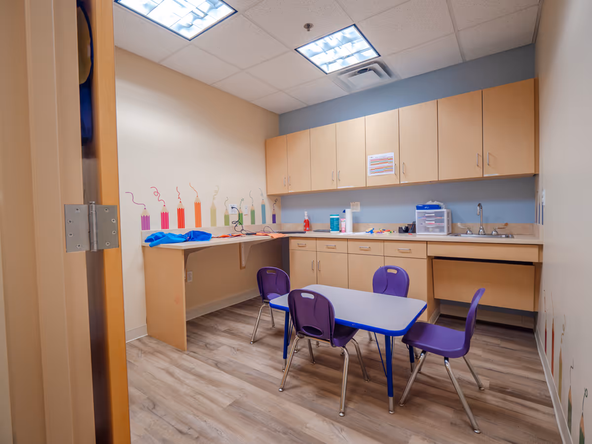 Interior classroom kitchenette with wooden cabinets, a sink, and a small table with purple chairs.