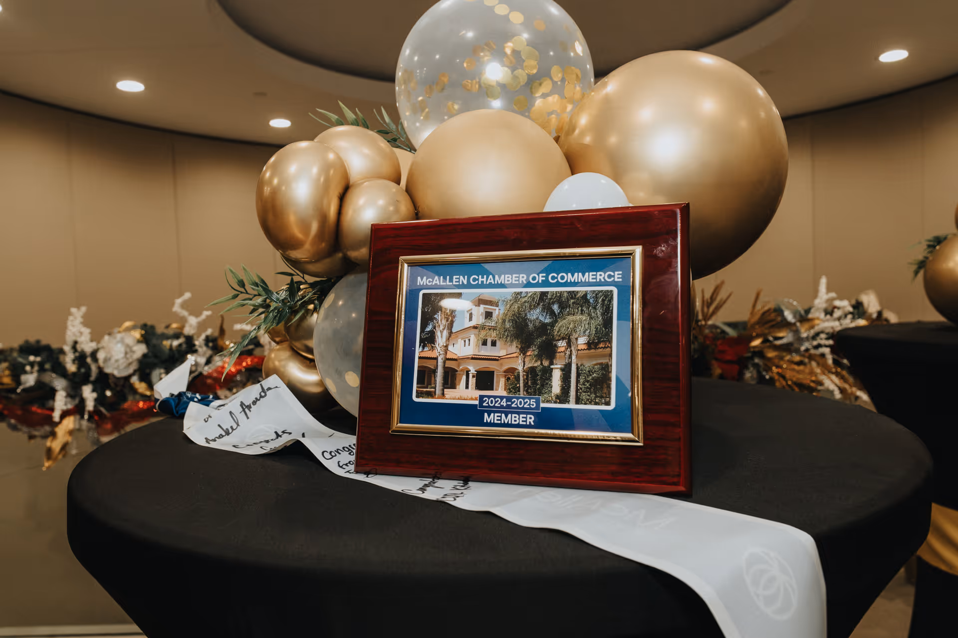 Award plaque on a table with gold balloons and a white ceremonial sash indoors.