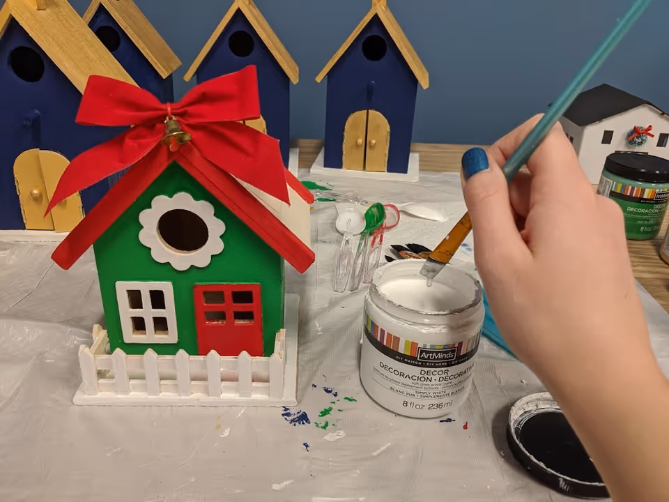 A hand with blue-painted nails paints a green decorative house model at a craft table.