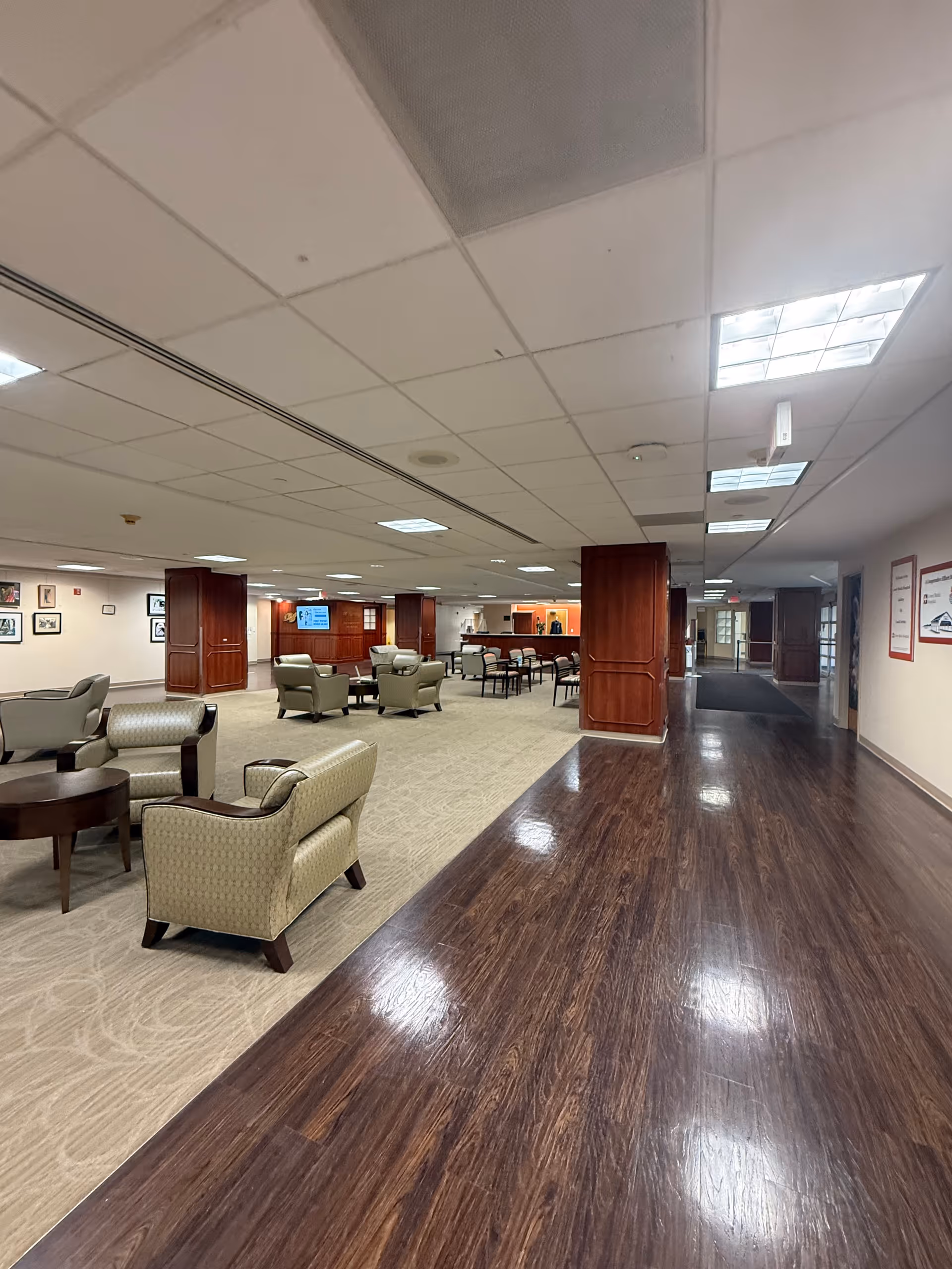 Interior lounge with beige chairs, tables, wood columns, and mixed carpet and dark wood flooring.