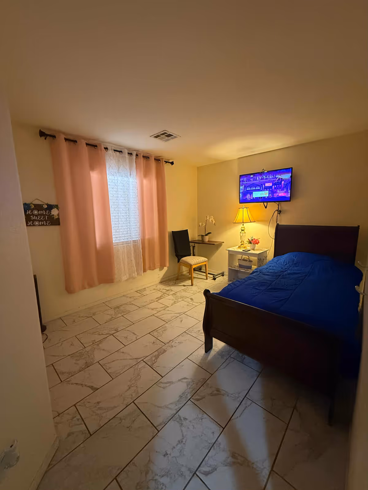 A small bedroom with a wooden bed, blue bedding, nightstand, desk, chair, and TV.