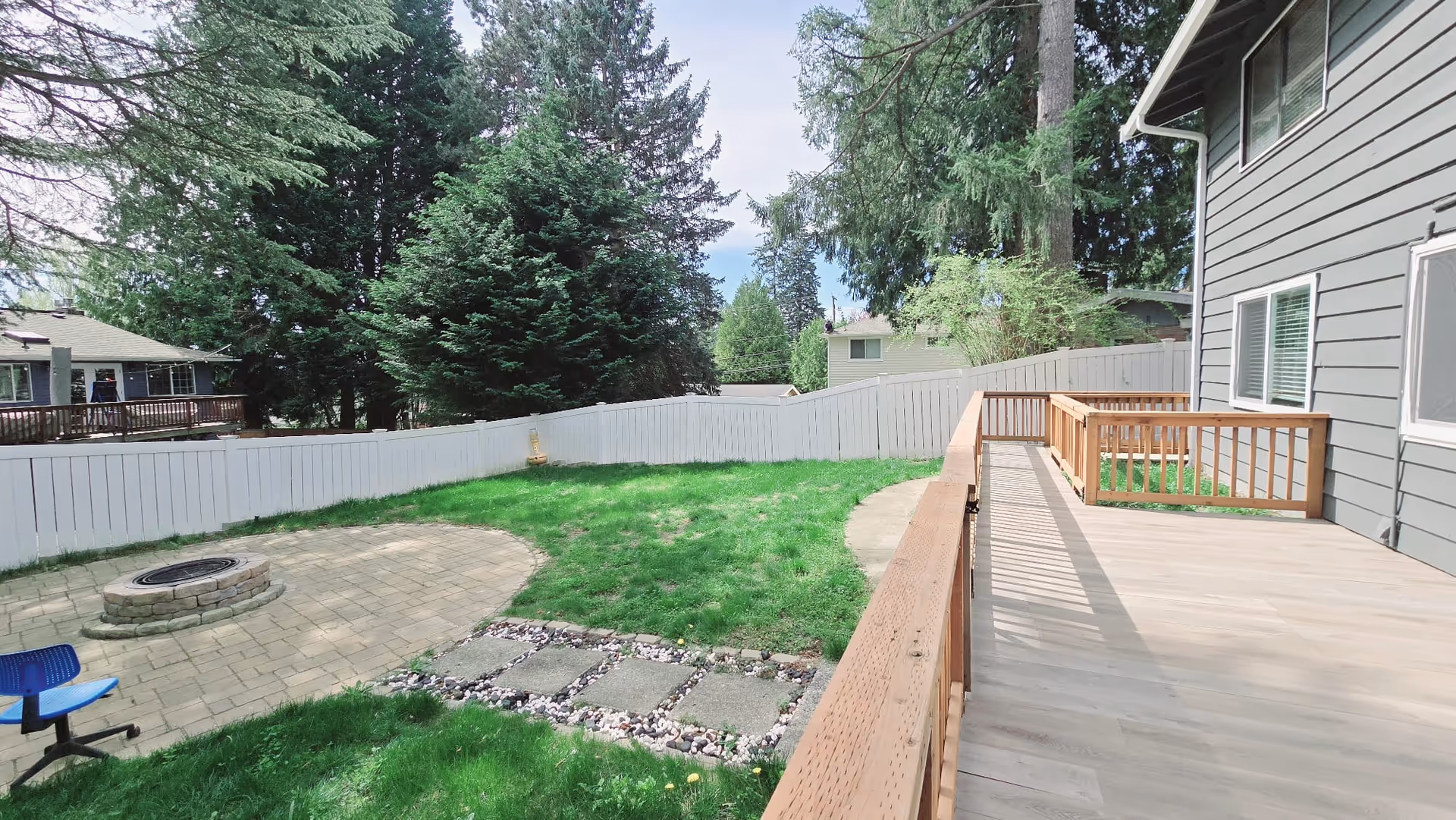 Backyard view of a gray house with a wooden deck, fence, fire pit, and lawn.