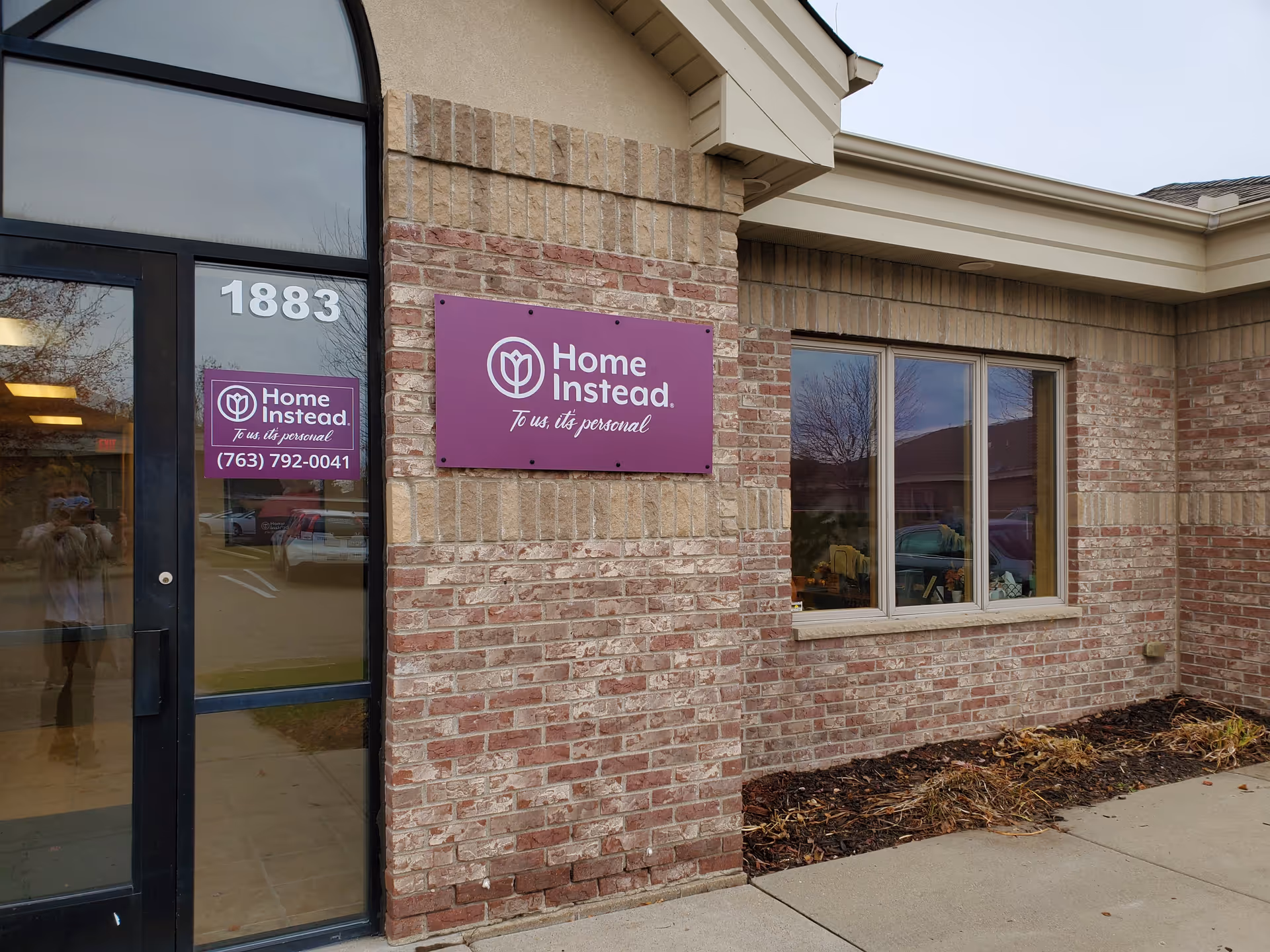 Front exterior of a Home Instead building with brick wall, door, window, and purple sign.