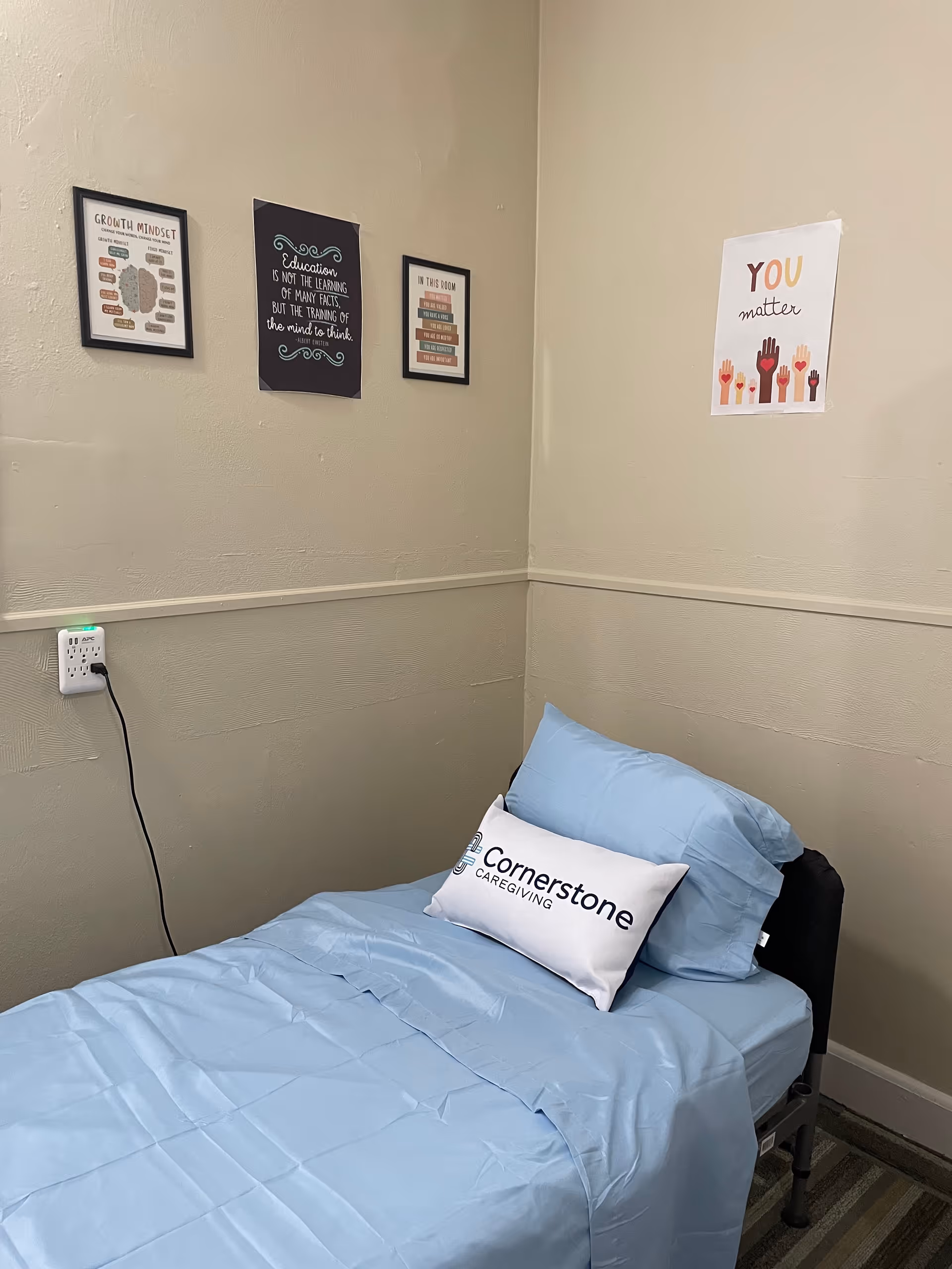 A small bedroom with blue bedding, framed posters, and a Cornerstone Caregiving pillow.