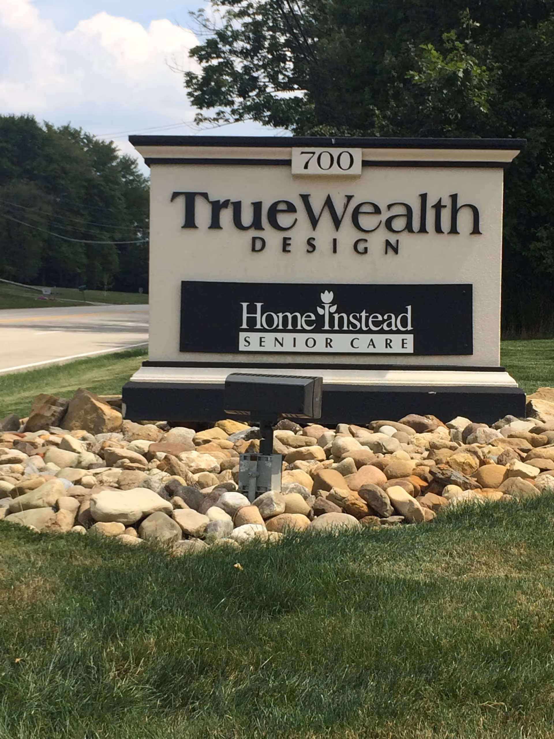 Outdoor sign shows TrueWealth Design and Home Instead Senior Care branding.