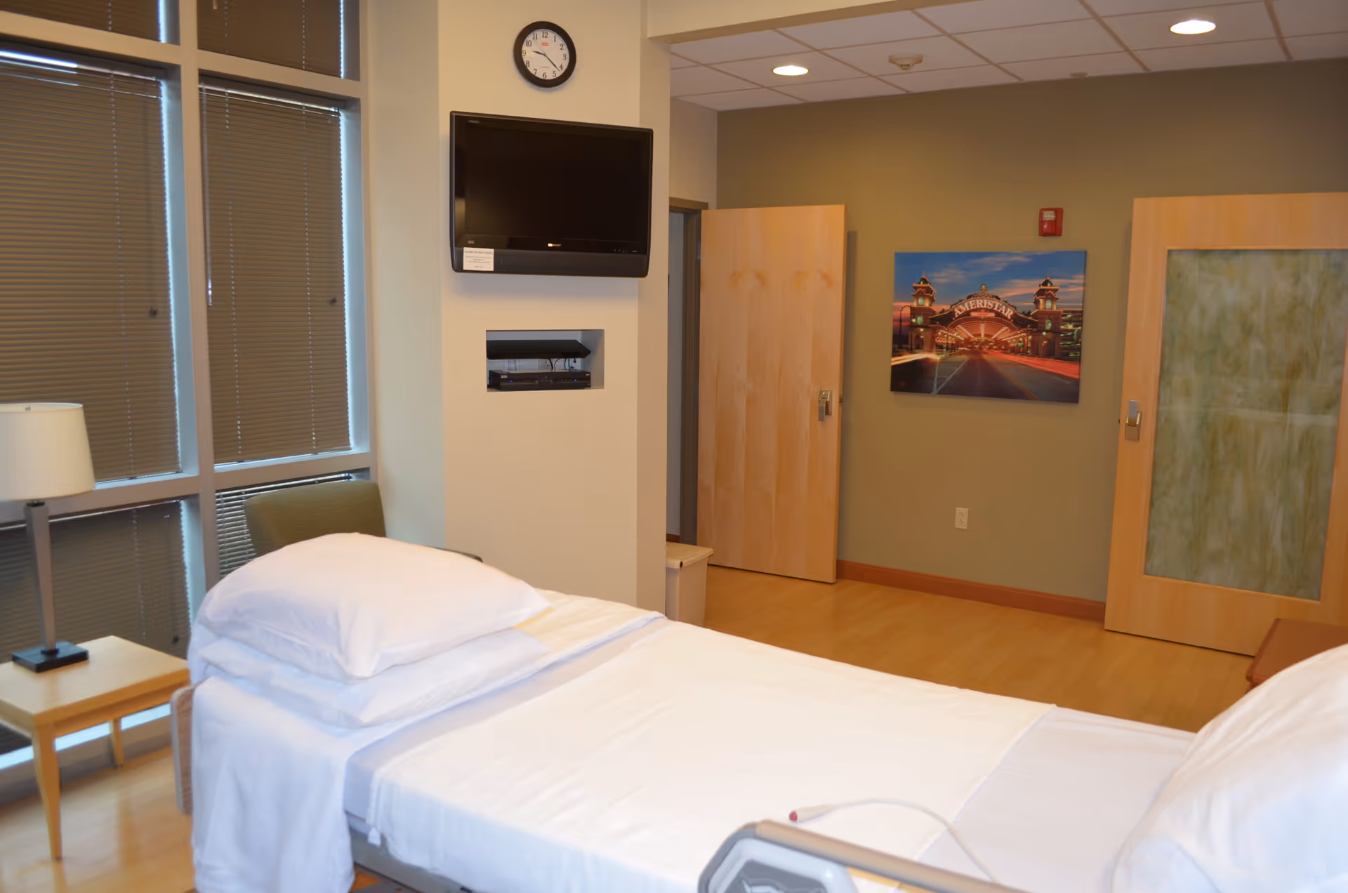 A hospital-style bed in a bright patient bedroom with blinds and wall-mounted TV.