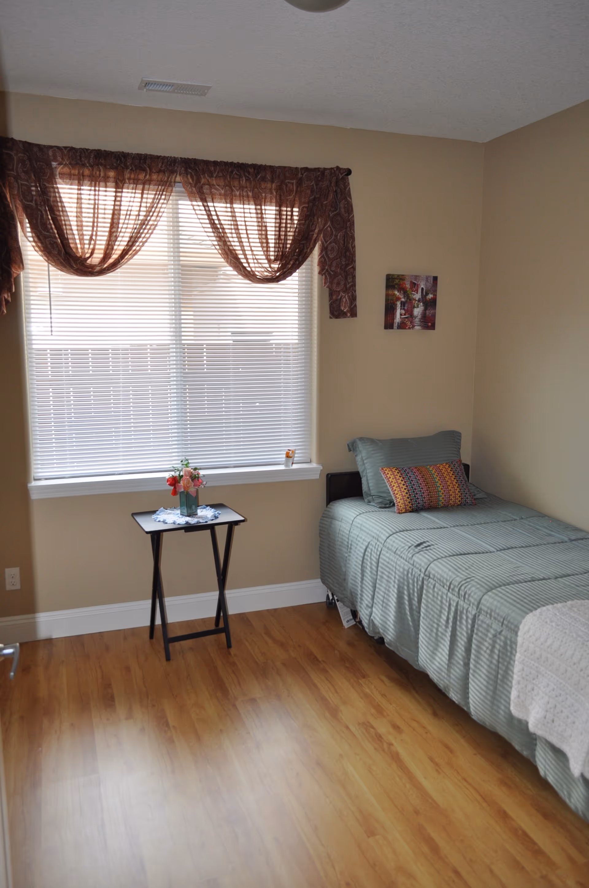 A tidy bedroom with a single bed, small table, and window with brown curtains.