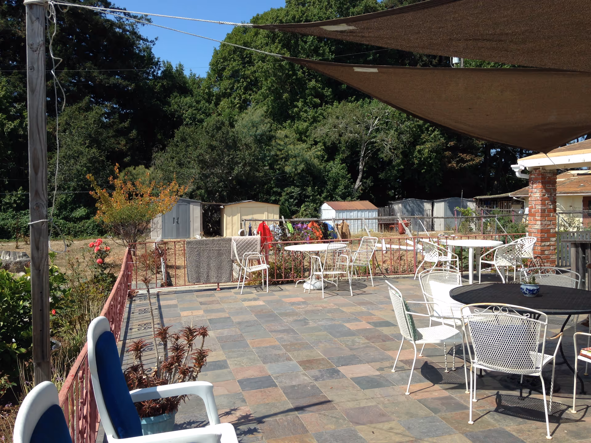 Outdoor tiled patio with white metal chairs and tables under shade sails, surrounded by greenery.