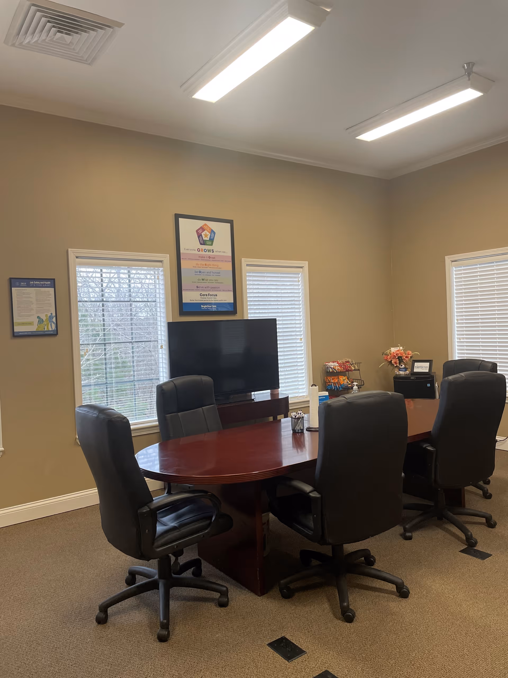 Interior conference room with an oval wooden table, six black rolling chairs, and a TV.