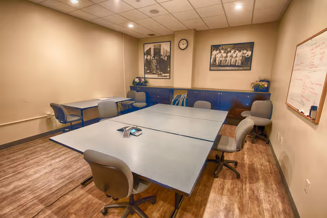 Conference room with large tables, chairs, whiteboard, and framed photos along blue cabinets.