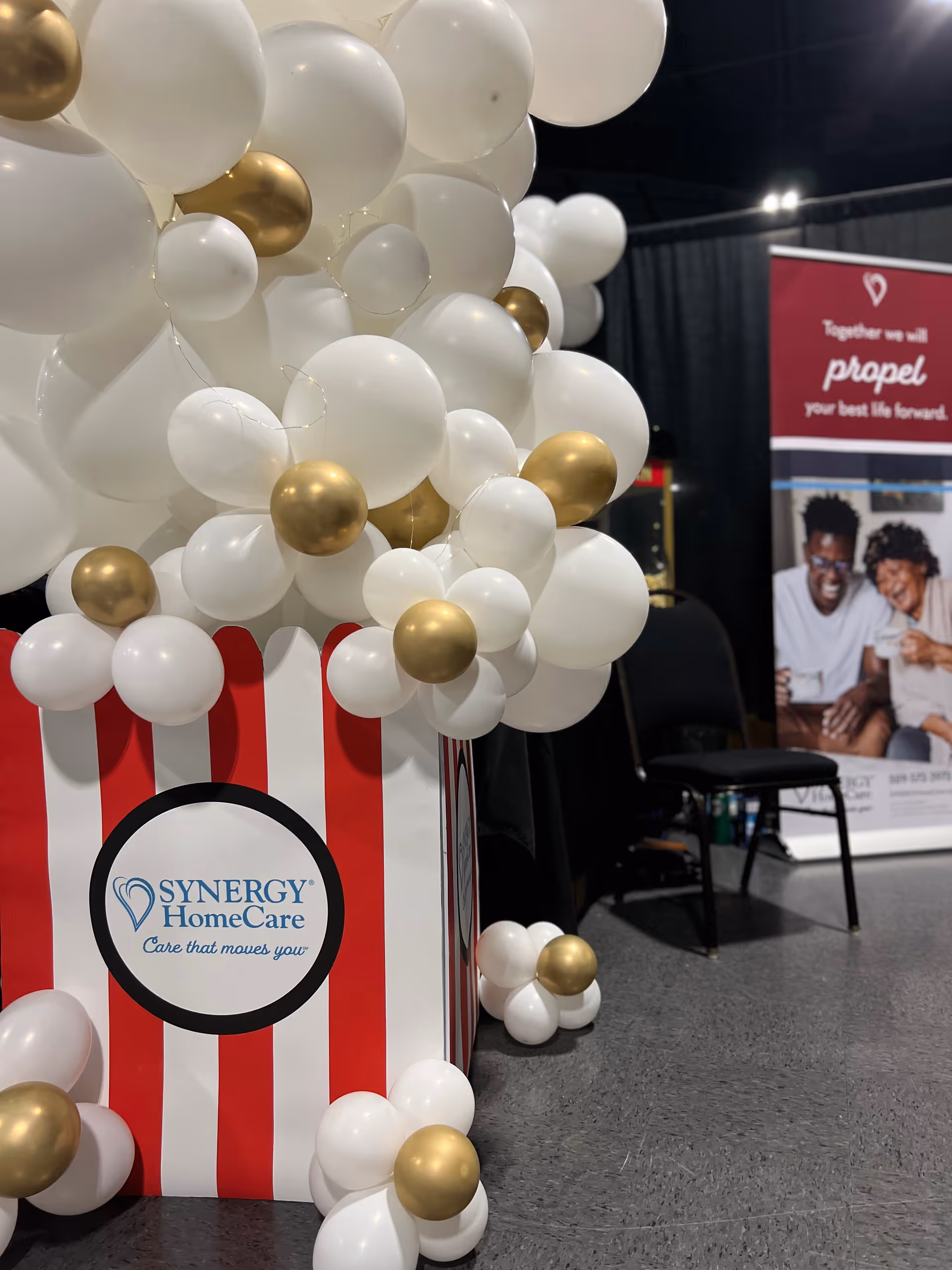 White and gold balloons surround a red-and-white Synergy HomeCare display indoors.