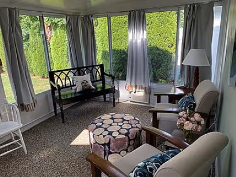Sunlit senior living room with multiple seats, a patterned ottoman, lamp, and floral decor.
