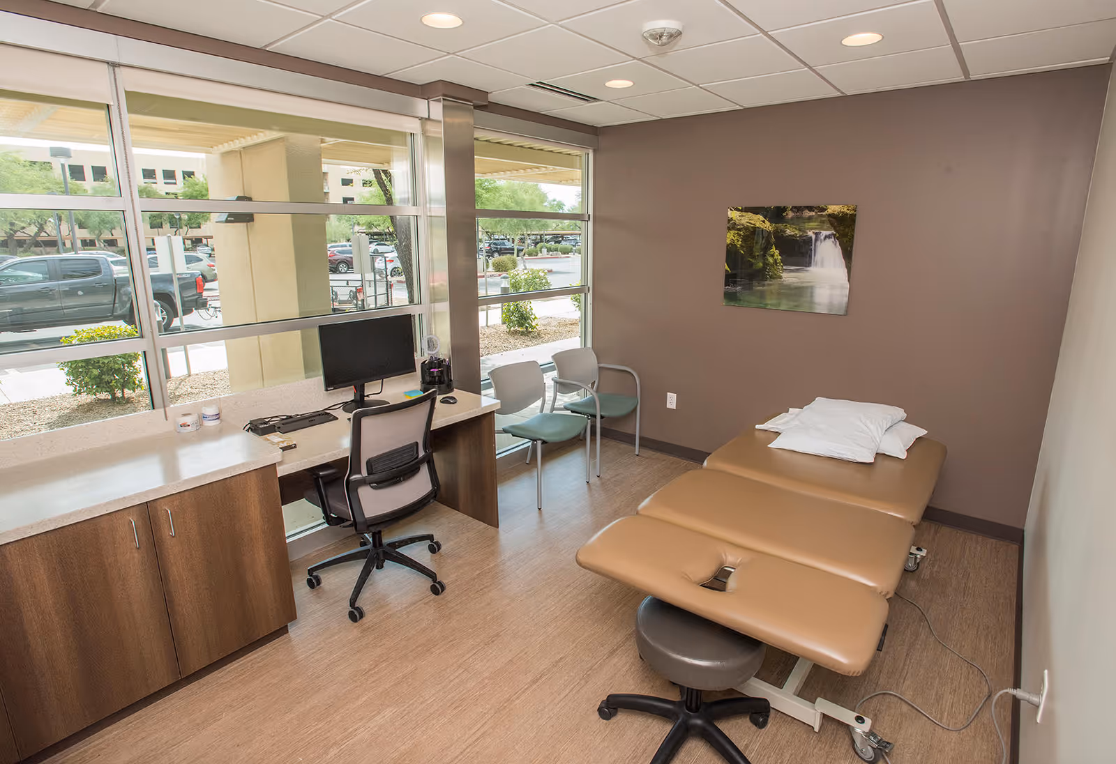 A therapy treatment room with desk, computer, and brown examination bed near windows.