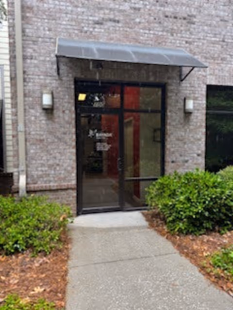 Brick building entrance with glass door, metal awning, and neatly trimmed shrubs