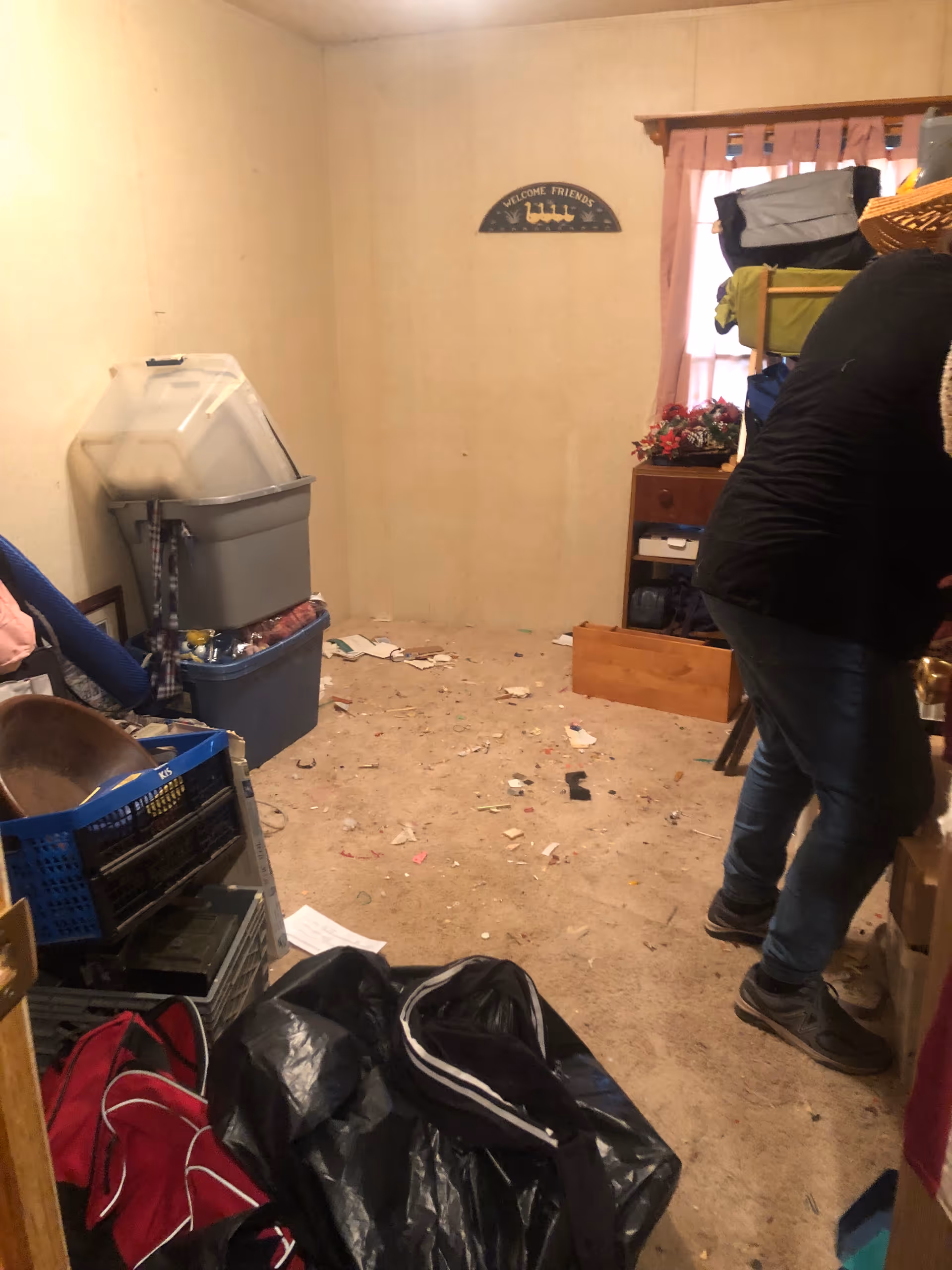A cluttered utility room with bins, trash bags, scattered papers, and a person nearby.