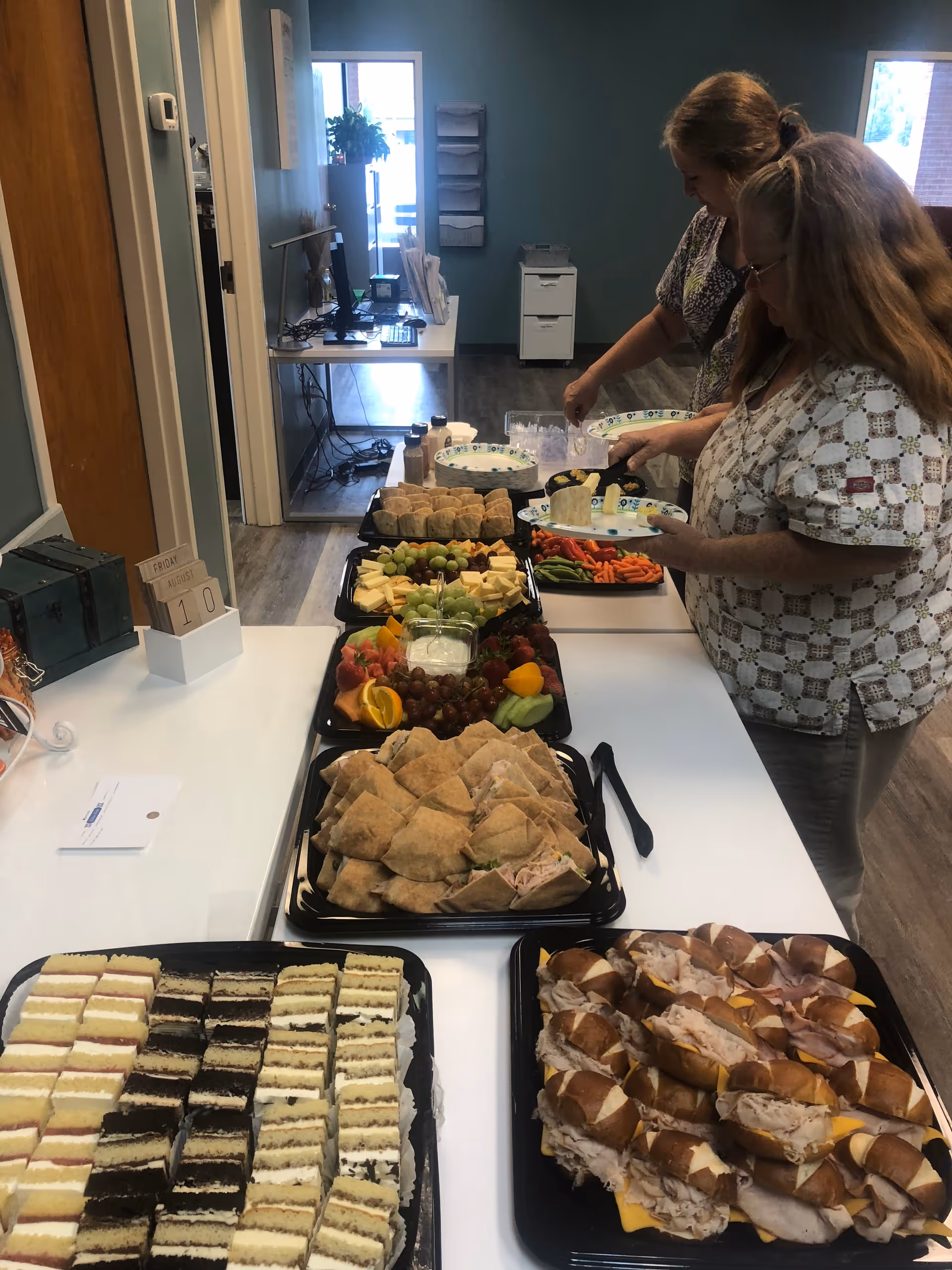 Two staff members arrange a buffet of fruit, pastries, and sandwiches for residents.
