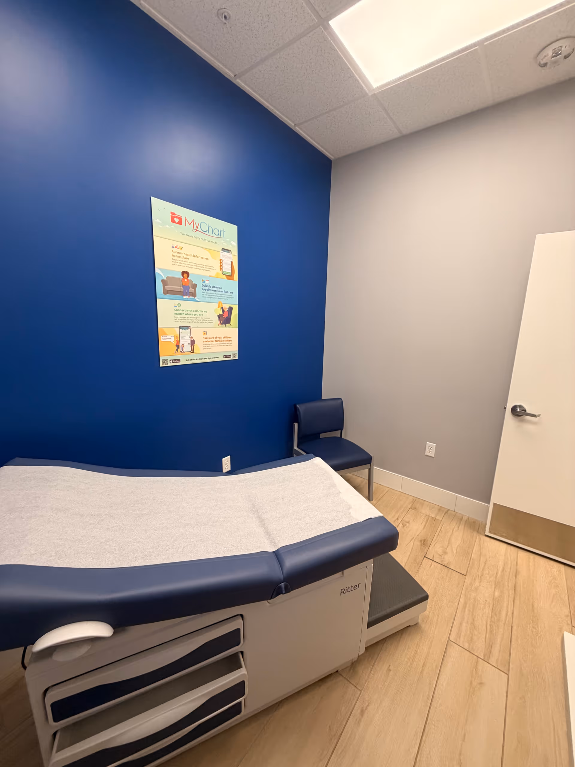 Blue-walled examination room with a padded medical bed, chair, and wall poster.