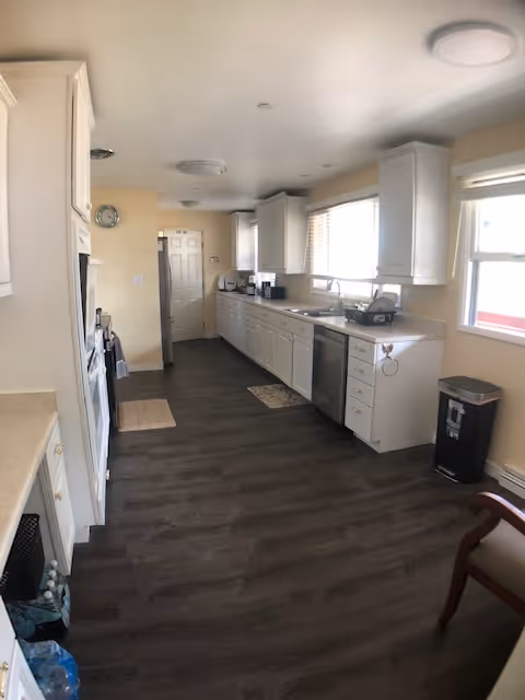 Bright kitchen in a senior living facility with white cabinets, appliances, and dark flooring.