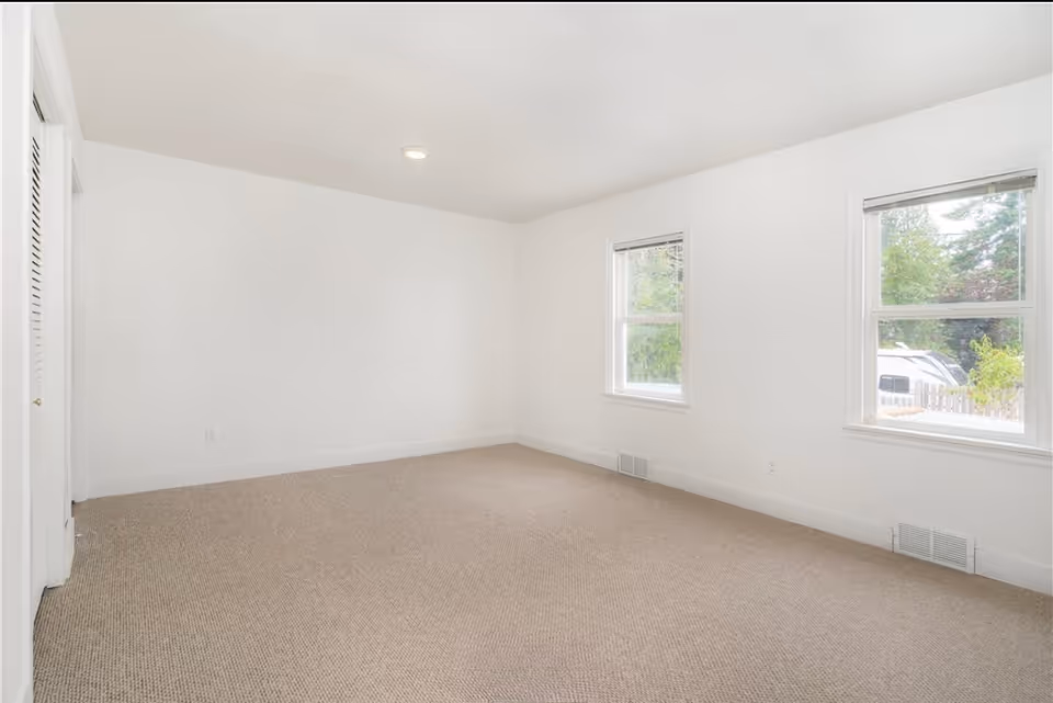 Bright, empty room with white walls, beige carpet, two windows, and a doorway.