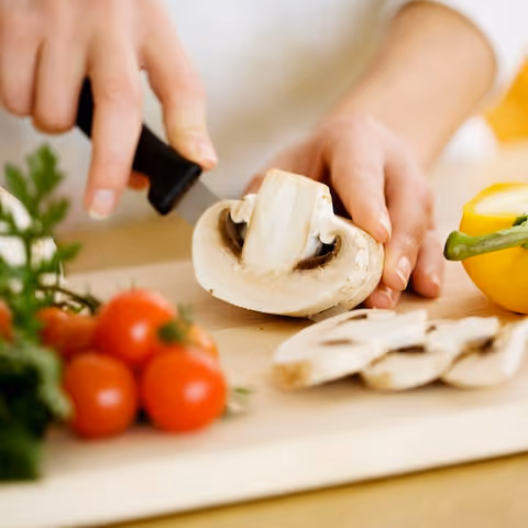 Hands chop mushrooms on a cutting board with tomatoes and peppers nearby