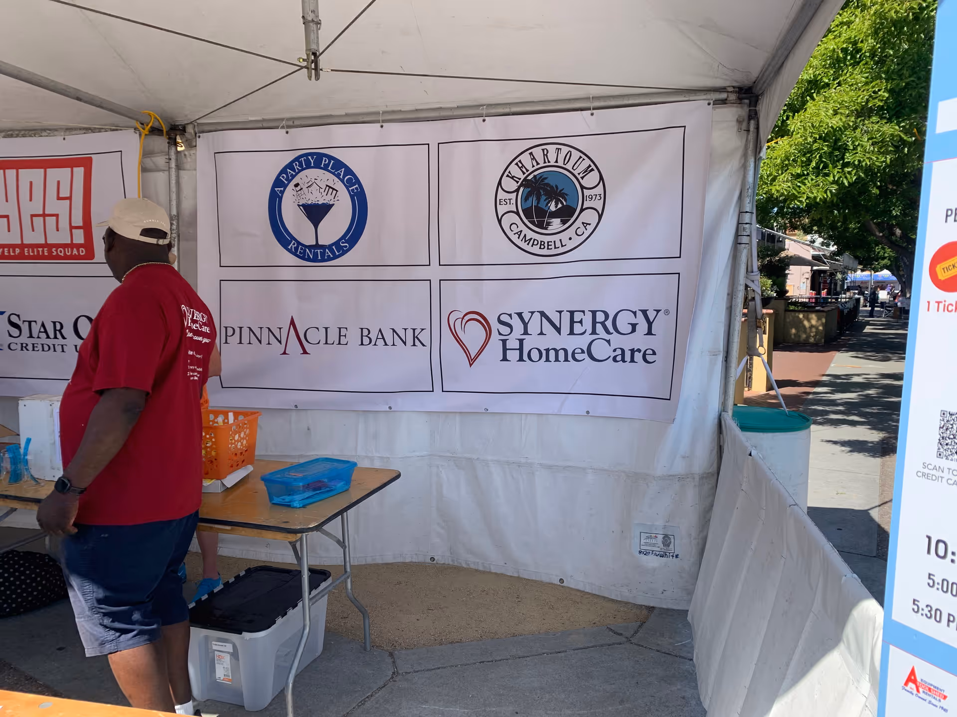 Man in red shirt stands beside a table under a tent with Synergy HomeCare banners.