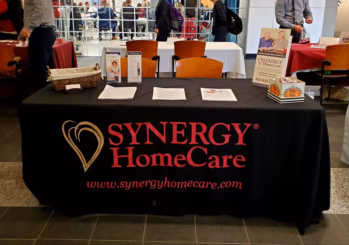 Indoor event booth table covered with Synergy HomeCare banner and promotional materials.