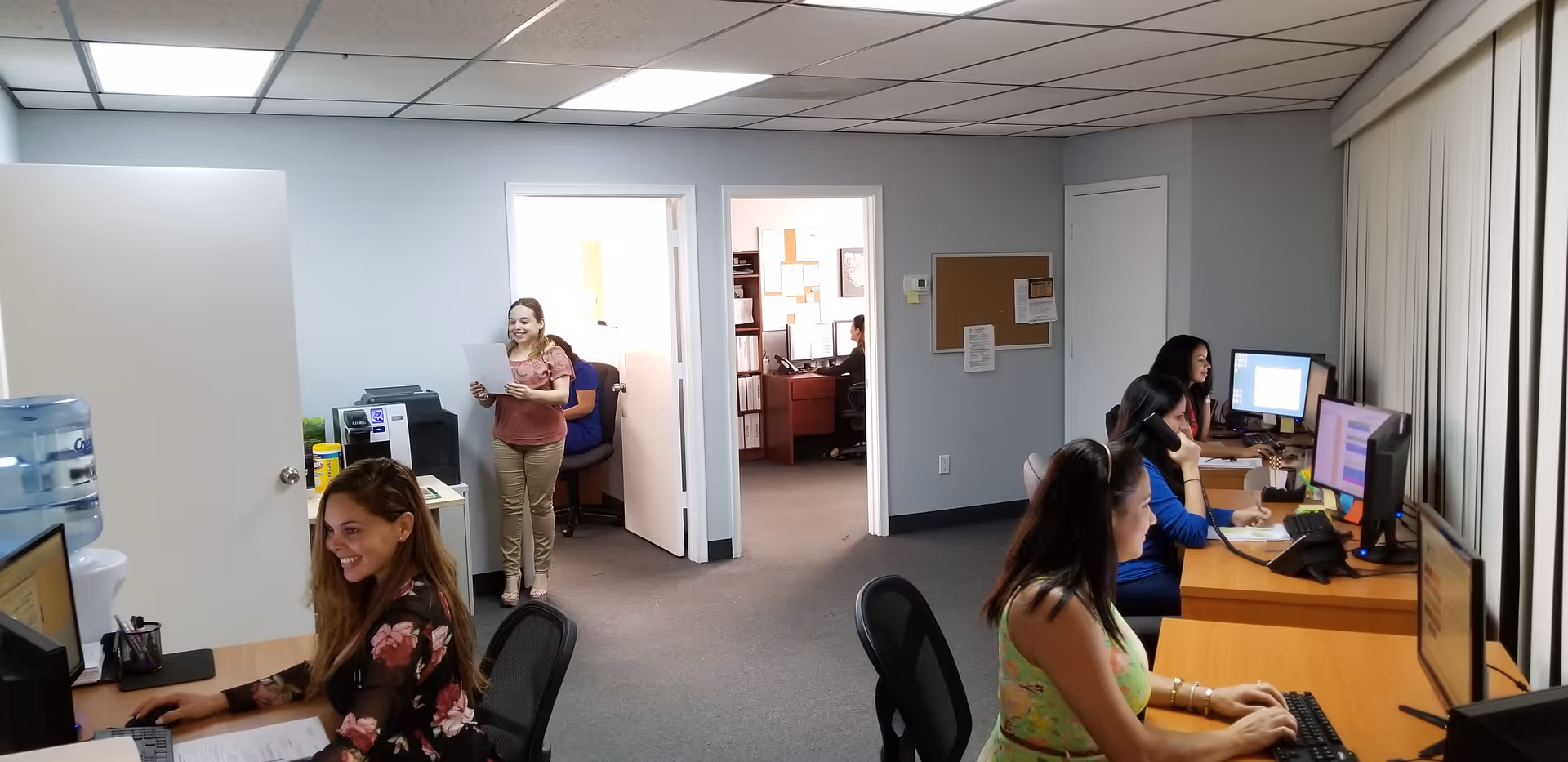 Office workspace with staff at desks, computers, and a standing woman holding papers.