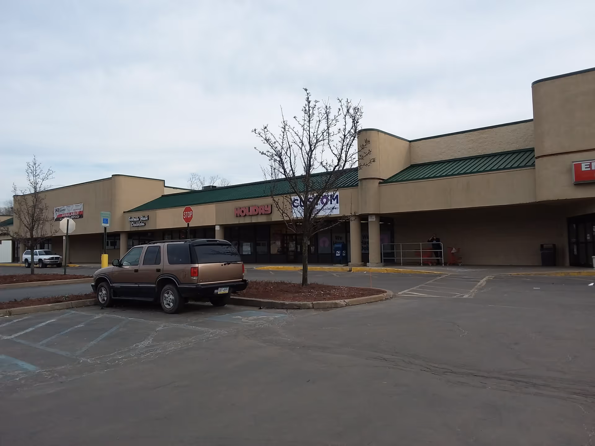 Exterior view of a strip mall with a parked SUV and leafless trees in front.