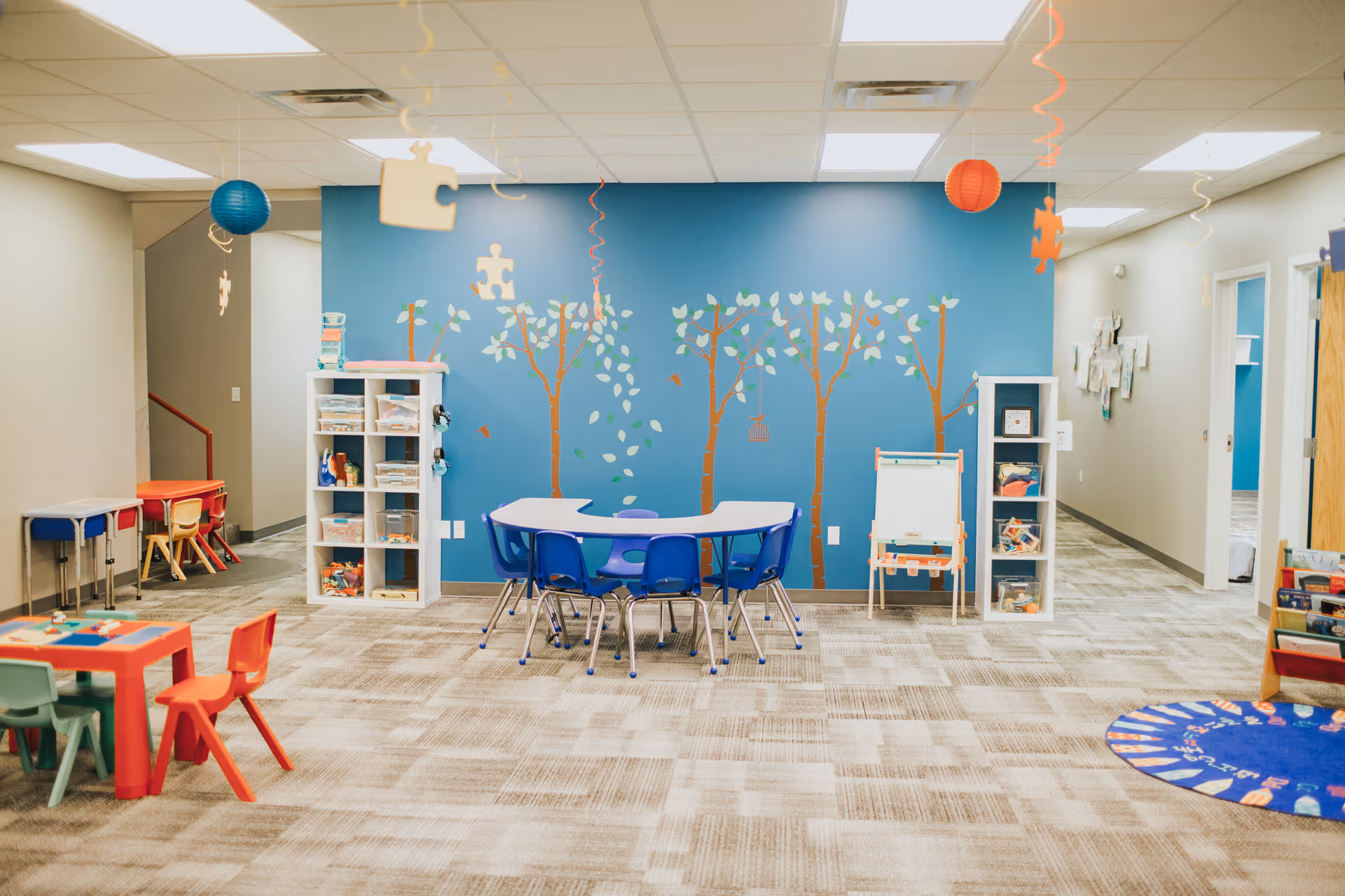 Colorful children's therapy playroom with blue tree mural, tables, chairs, and shelves.