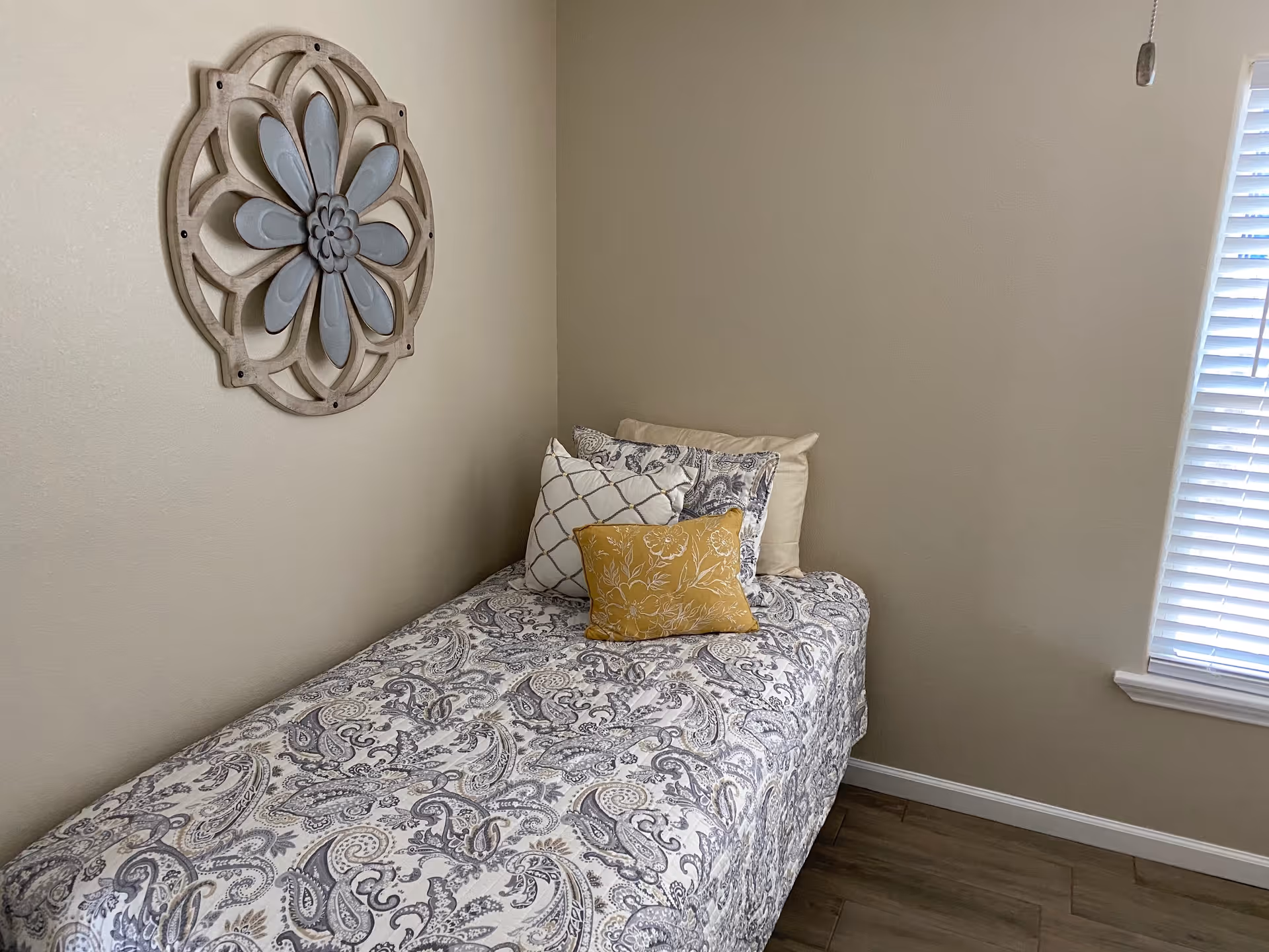 Single bed with paisley-patterned bedding, decorative wall piece, and a window with blinds.