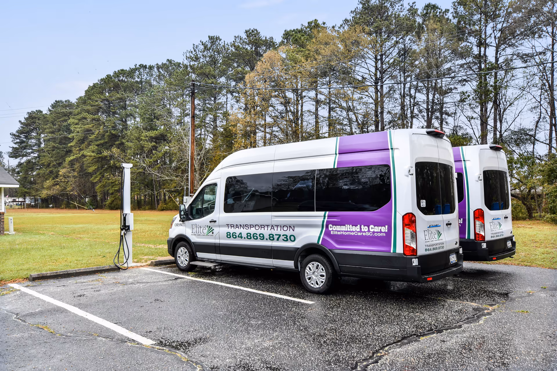 Two Elite Home Care vans parked in a lot beside a grassy field with trees.