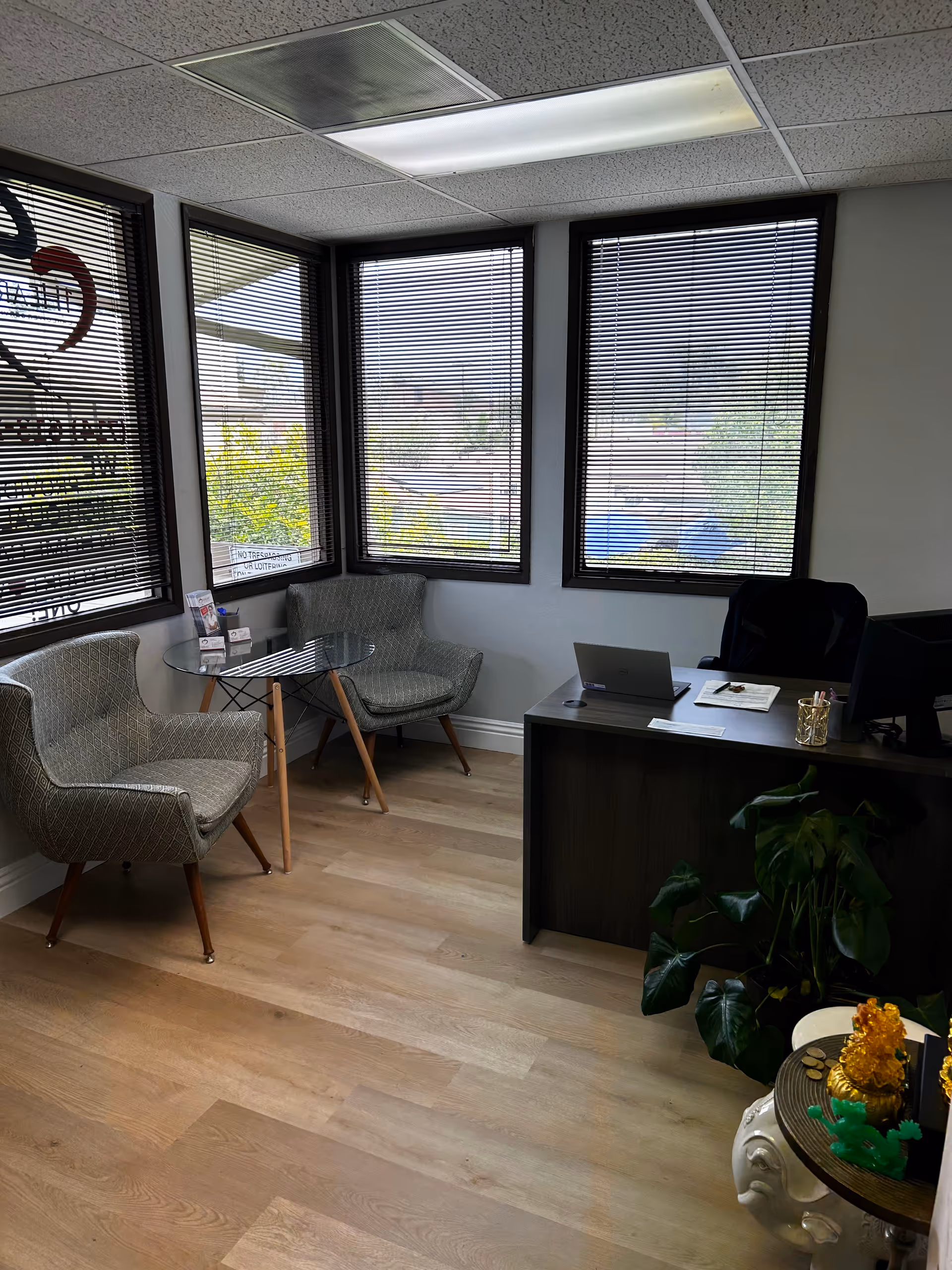 A small reception area with glass table, three chairs, a desk with laptop, and blinds.