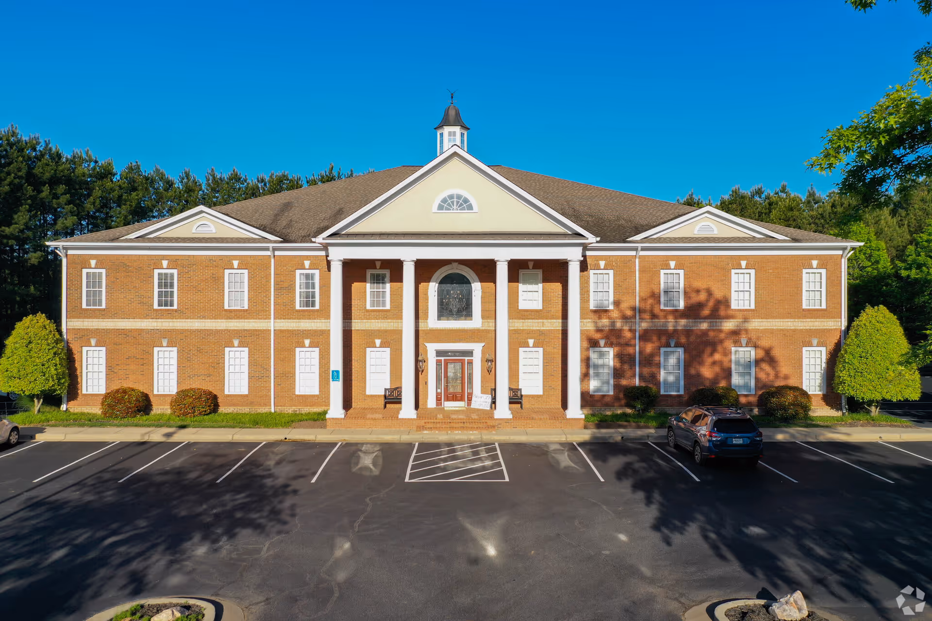 Brick building with columns and a curved entrance, parking lot and surrounding trees.