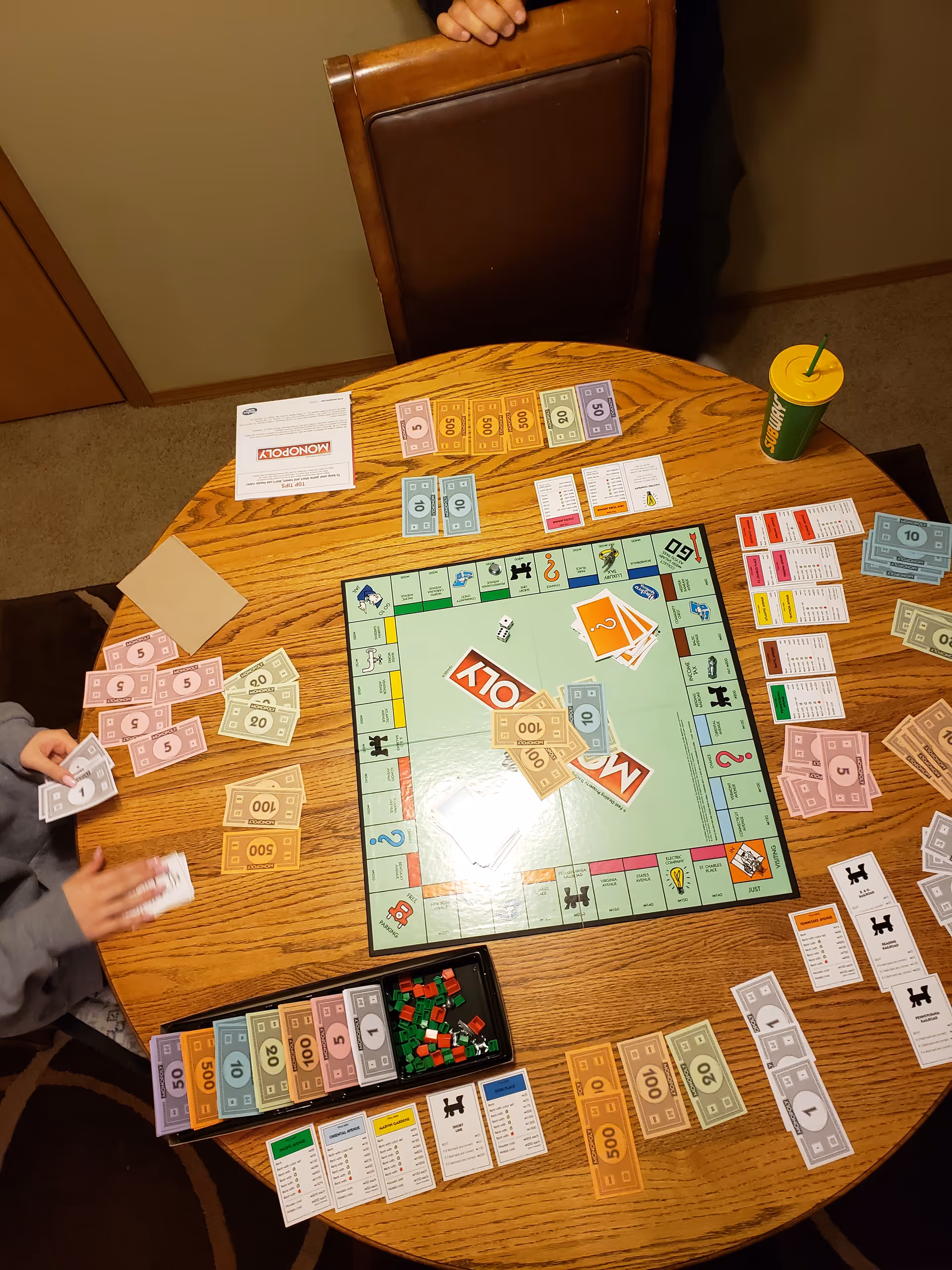 A round wooden dining table with Monopoly money, cards, game pieces, and a drink.