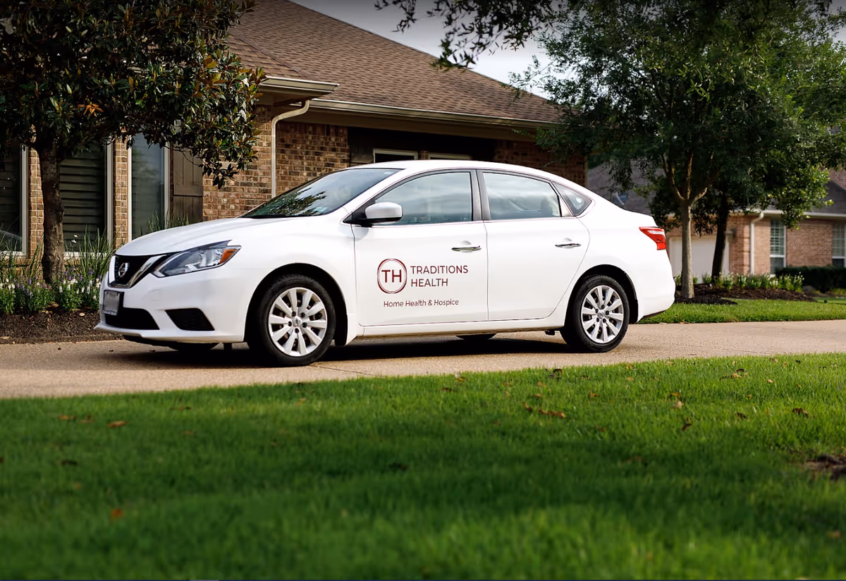 A white Traditions Health car parked in front of a brick hospice facility.
