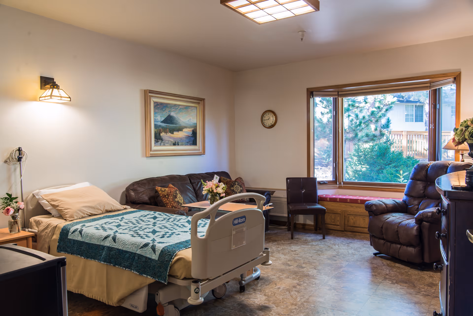 Senior living room with a hospital bed, leather sofa, chairs, and a large window.