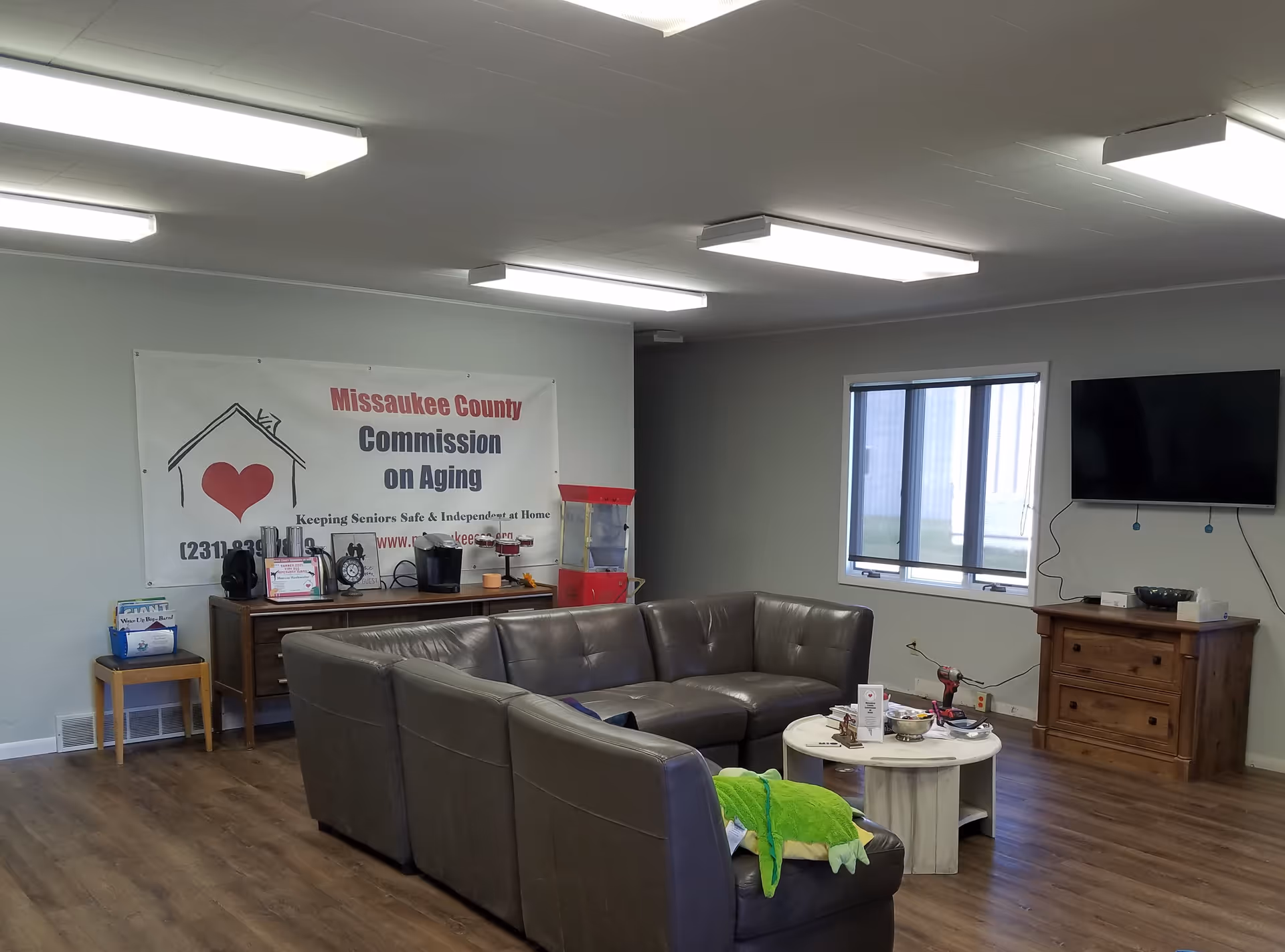 Lounge area with sectional, coffee table, banner reading Missaukee County Commission on Aging, and a TV.