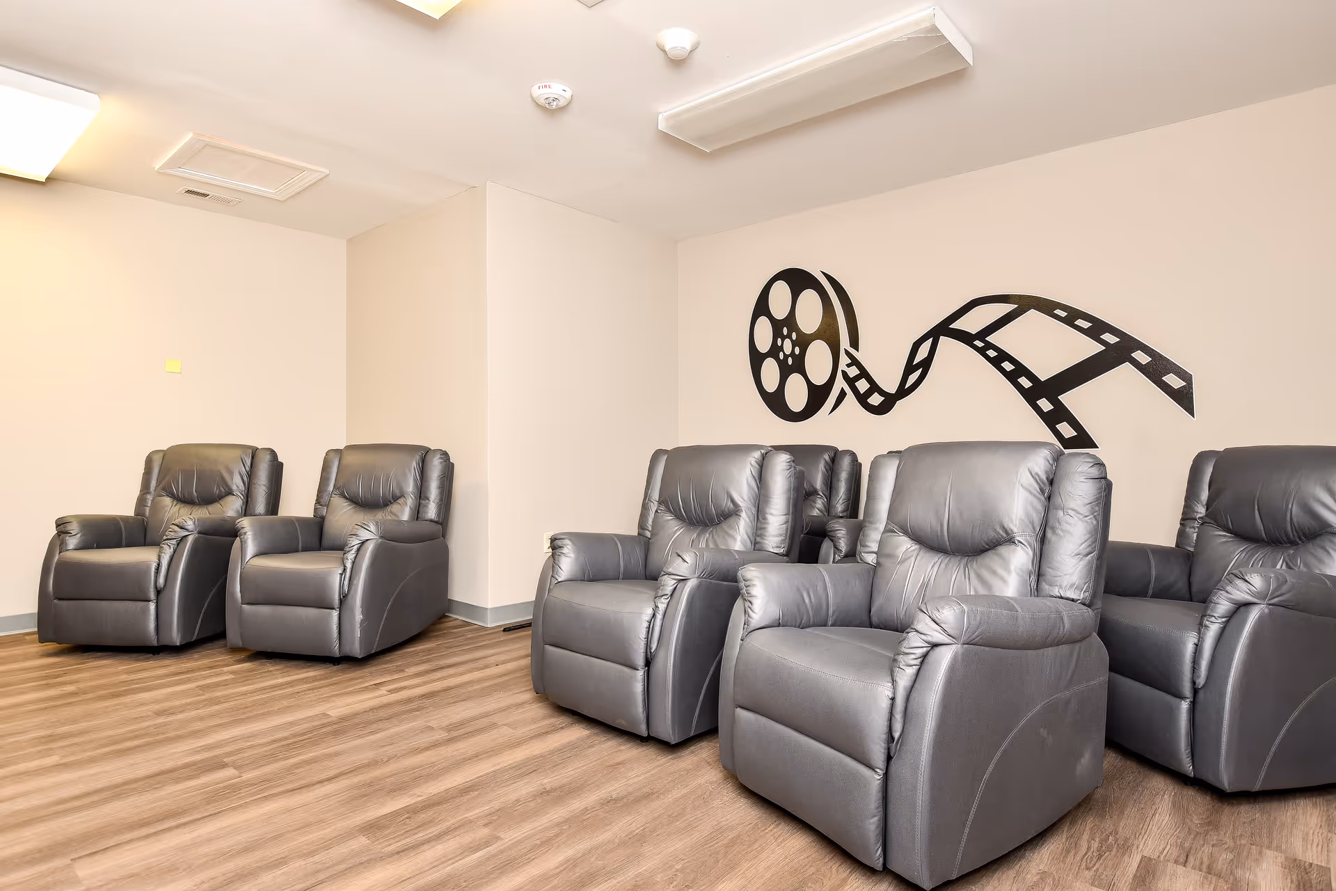 A modern senior living lounge with gray recliners, wood flooring, and film-wall art.