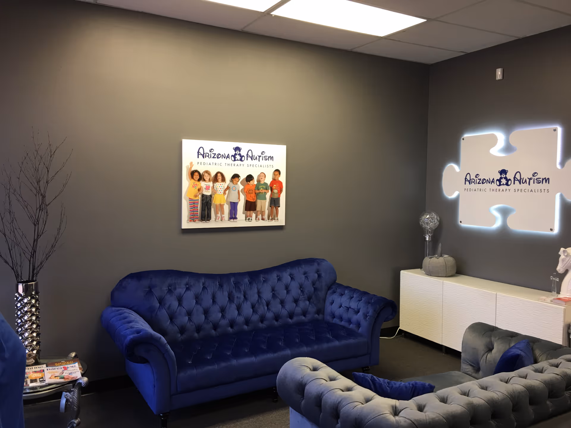 Gray walls surround a lounge with blue tufted sofas, white cabinets, and a puzzle-piece logo.