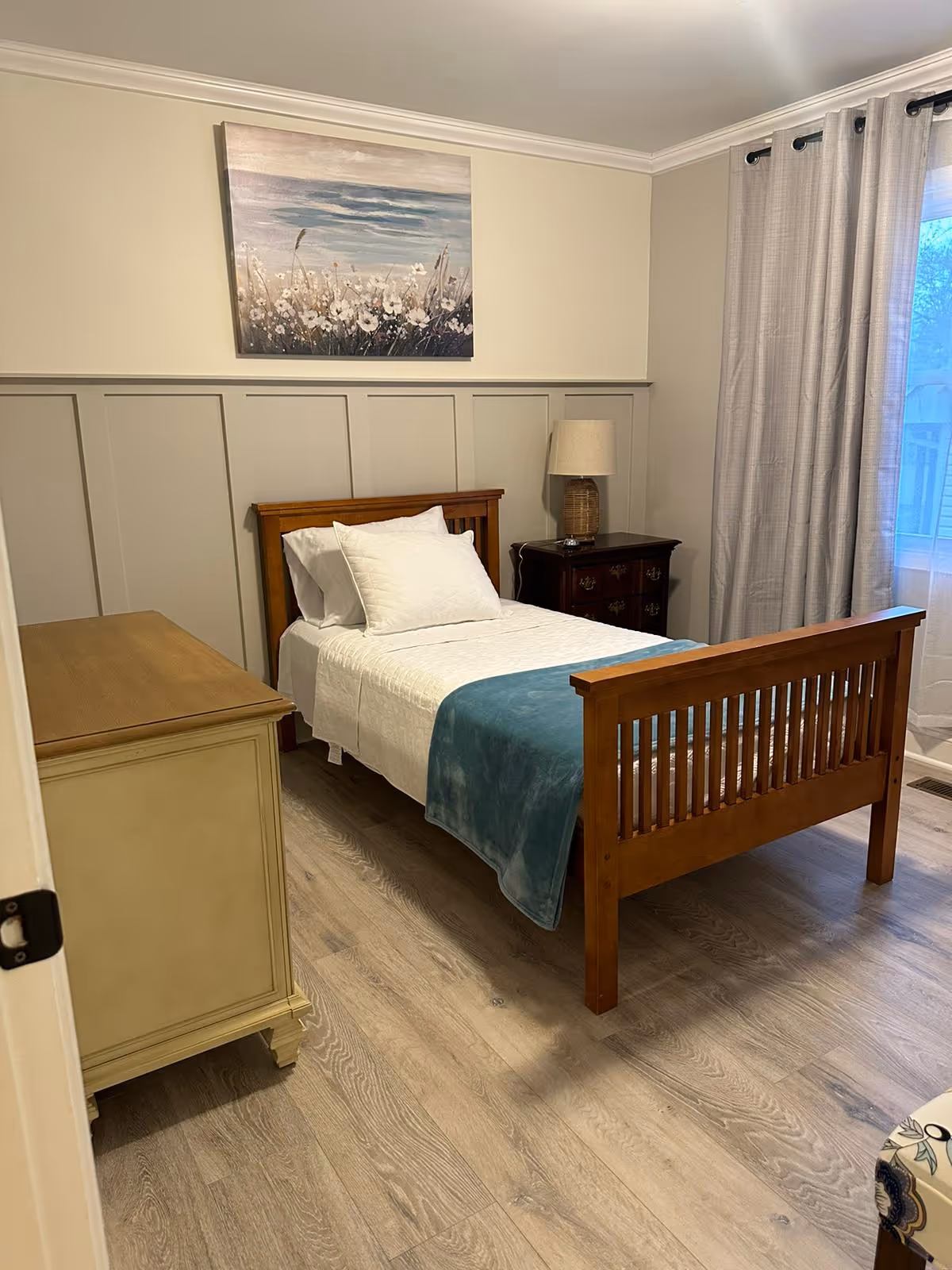 A tidy bedroom with wooden bed, dresser, nightstand, lamp, and light curtains.