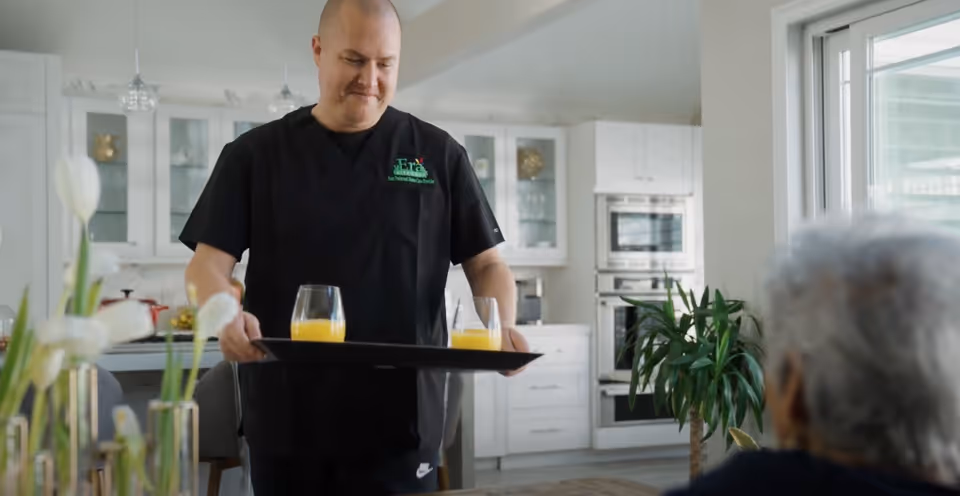 A server in black scrubs carries a tray with two orange juice glasses in a kitchen