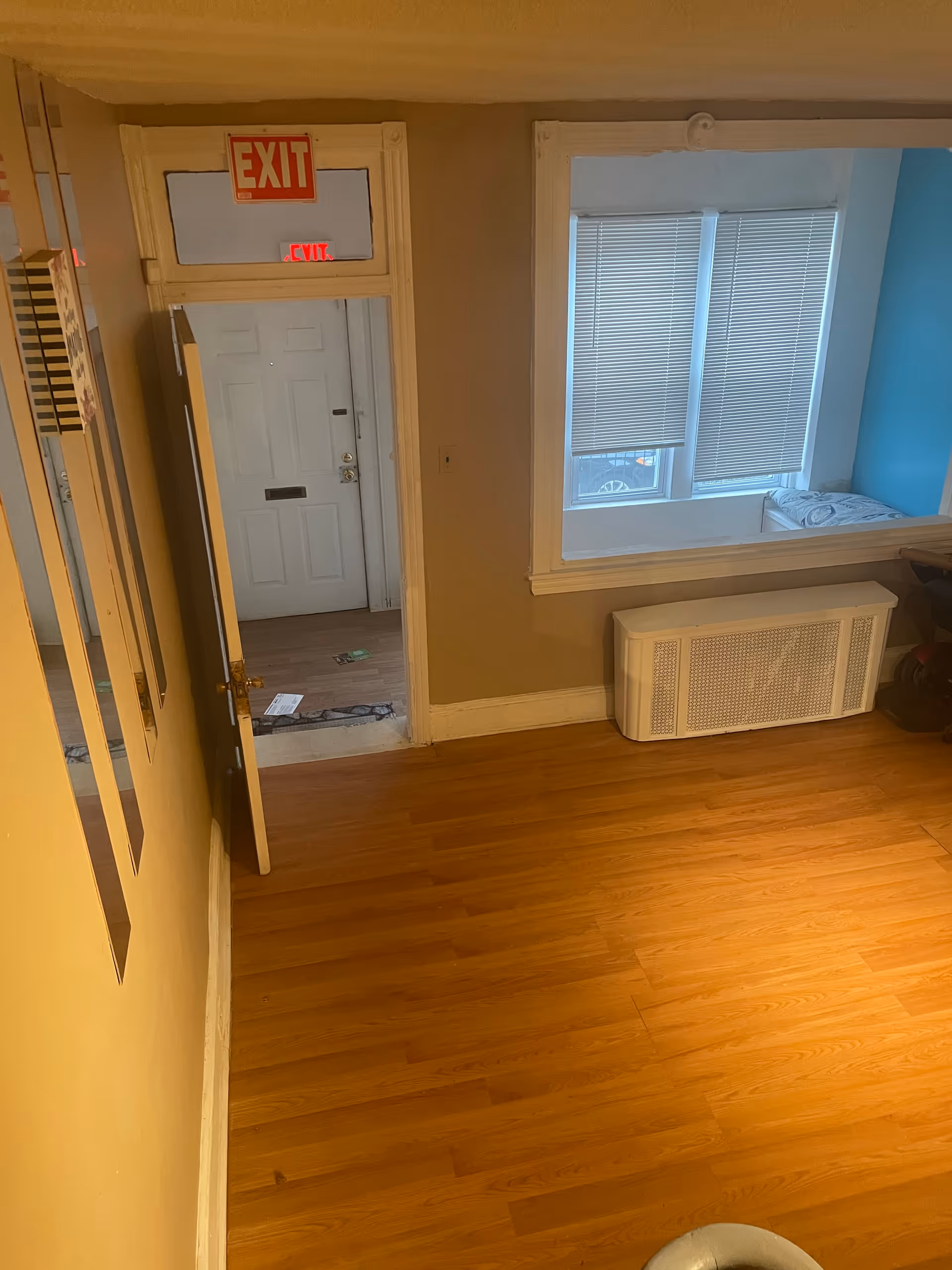 Interior hallway with wood flooring, exit door, window blinds, and radiator cover in a care home.